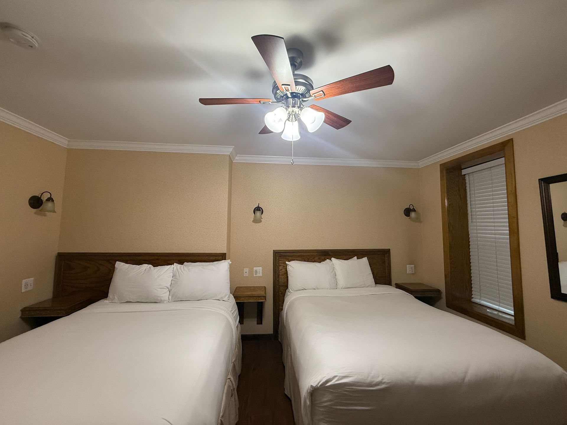 Two beds in a beige-walled room. A ceiling fan, nightstands, and a window are visible.