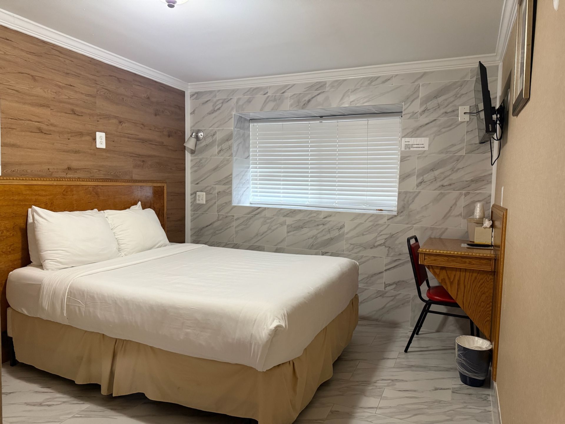 A hotel room with a bed, desk, and window. The walls are tiled and paneled, and the room has a simple, functional design.