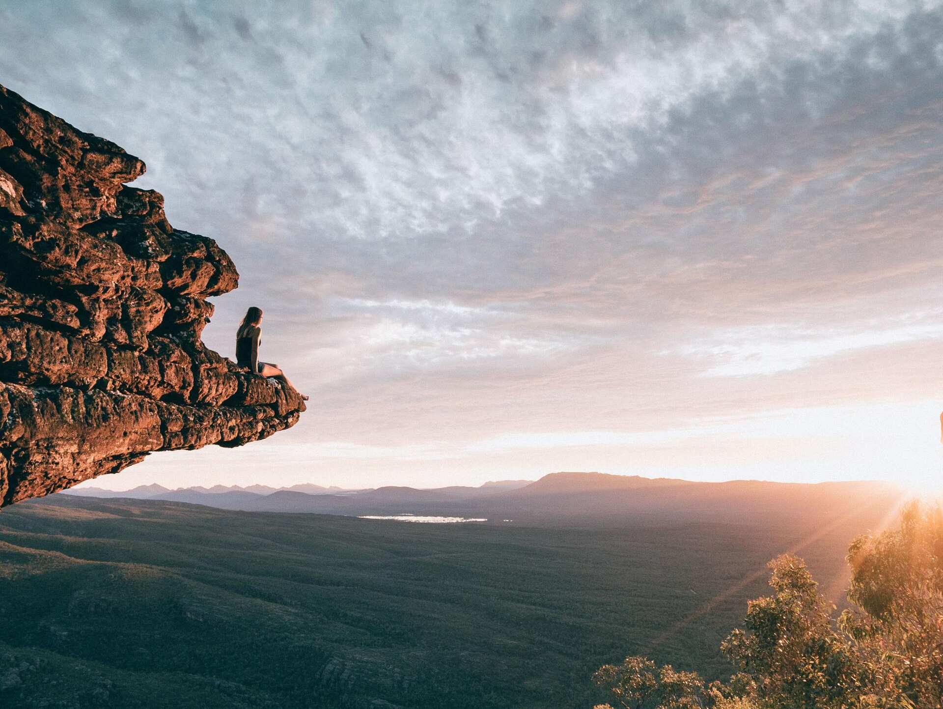 A person is sitting on a rock overlooking a valley at sunset.