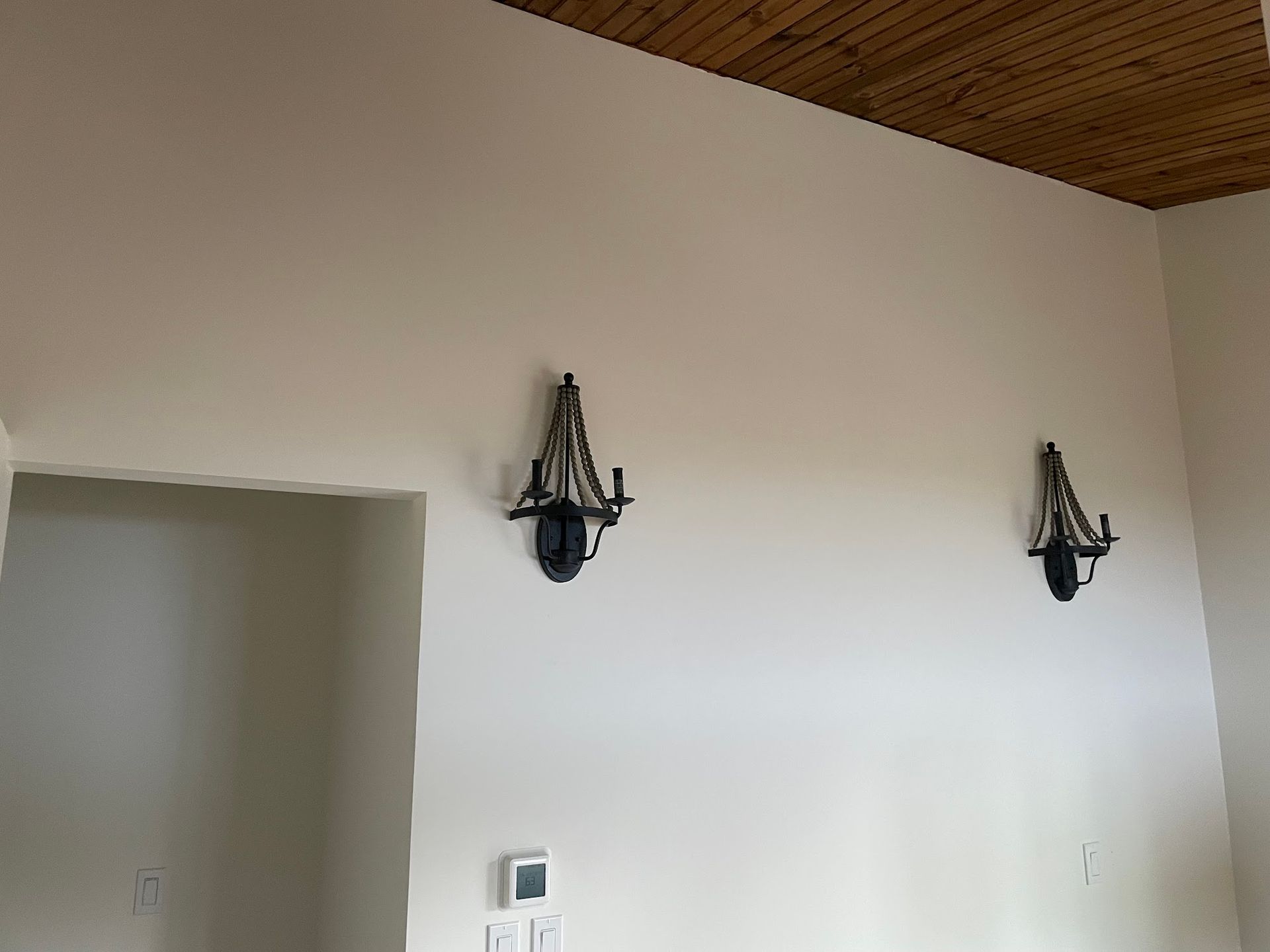 Two black sconces on a light beige wall, near a doorway and a light switch.  A wood ceiling is above.