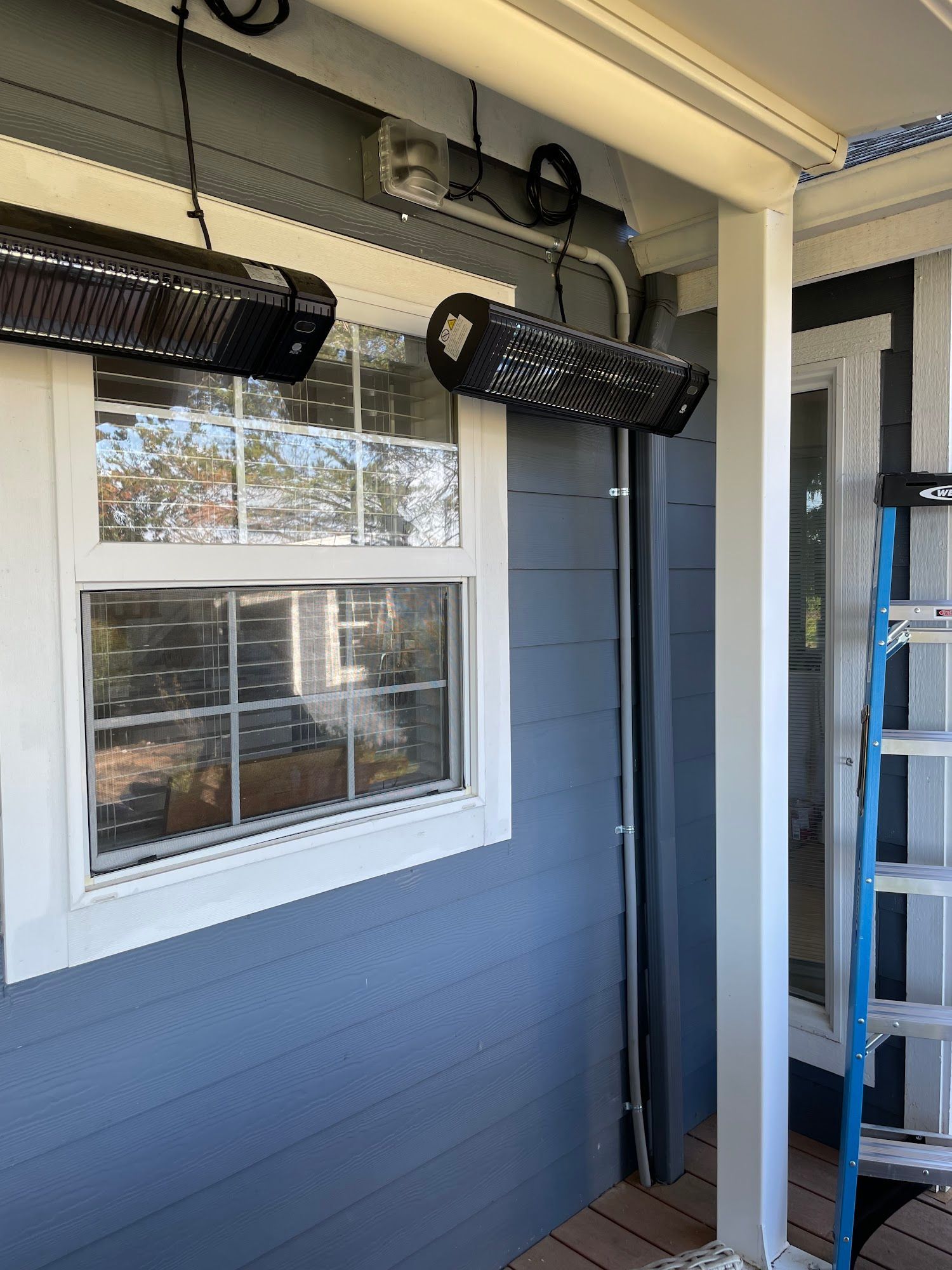 Two black infrared heaters mounted on a blue house's porch, next to a window and a white column.