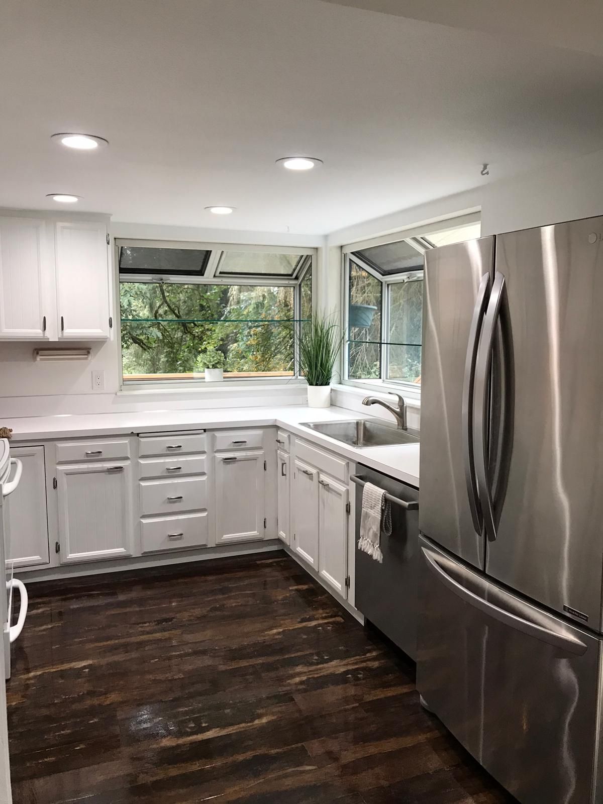 White kitchen with stainless steel appliances, dark wood floor, and a window overlooking greenery.