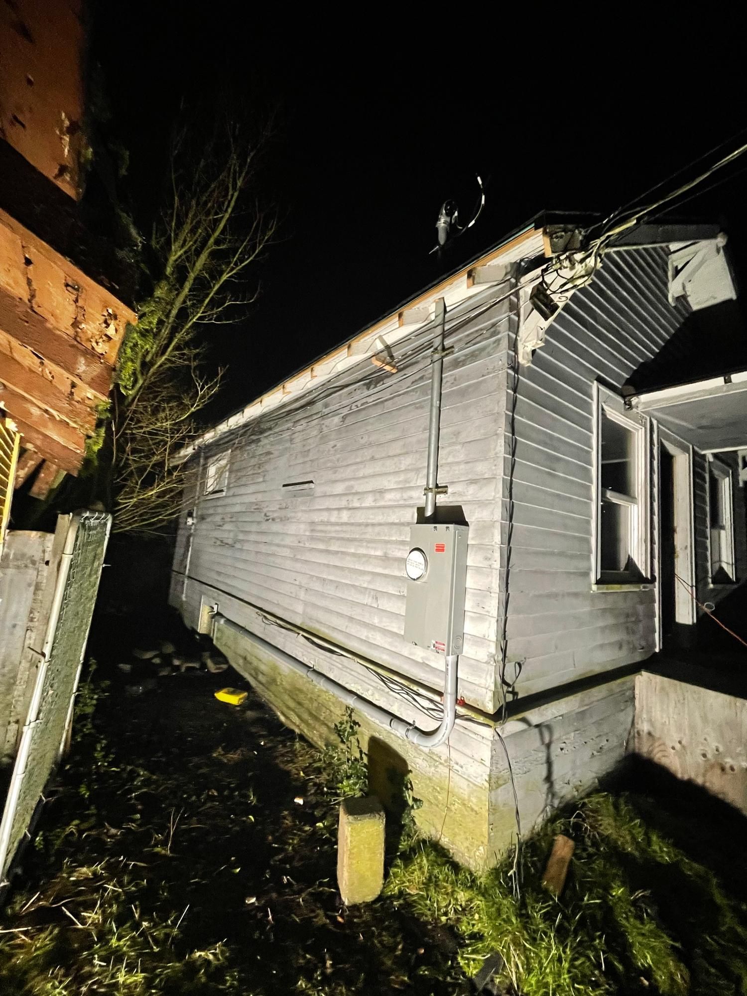 Side of a house at night, with electrical box and wires visible. Overcast sky.