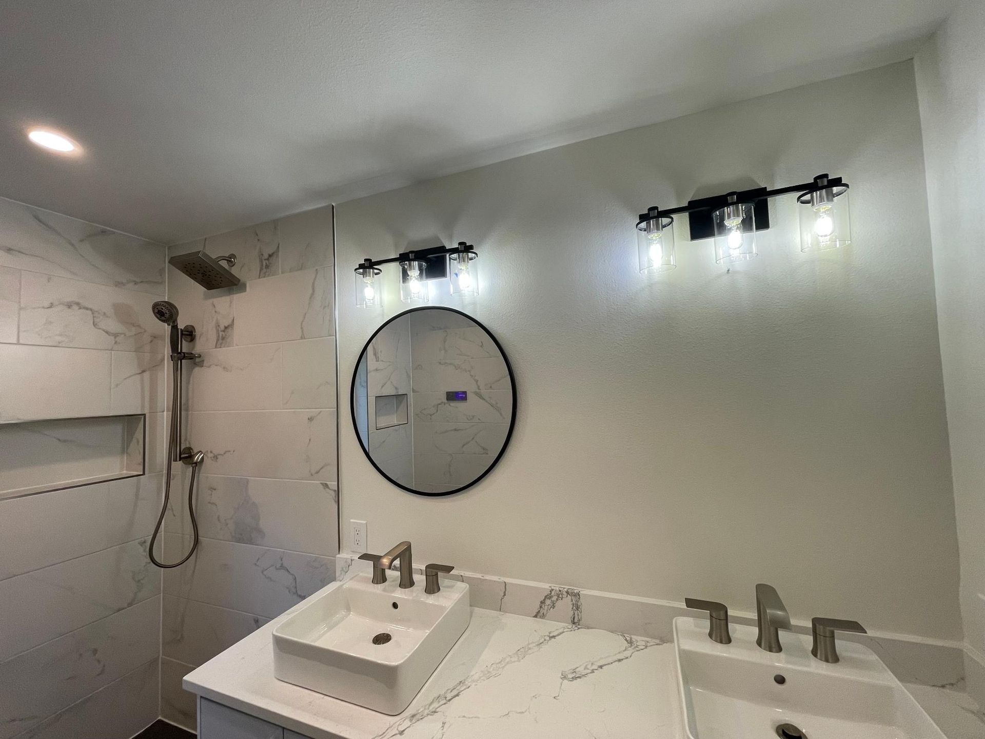 Bathroom with marble tile shower, double vanity with sinks, round mirror, and black light fixtures.