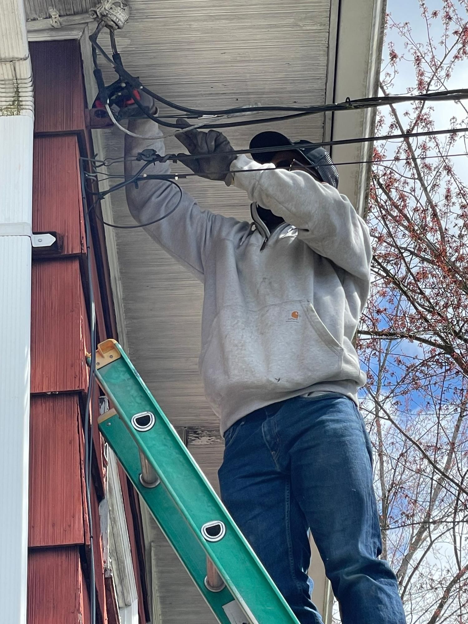 Person on a ladder working on overhead wires near a building's roof.