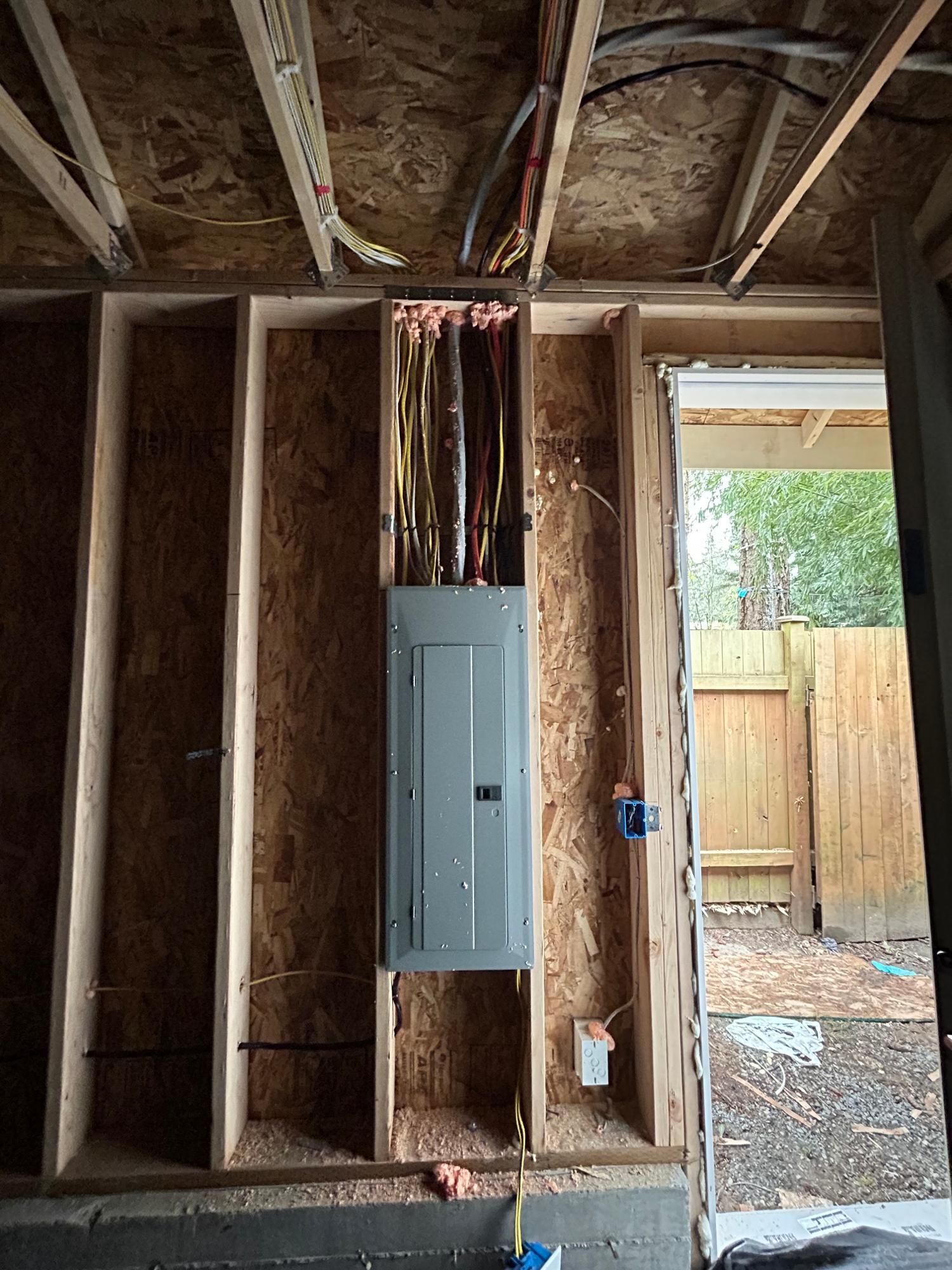 Electrical panel installed in a wooden wall, with wires running above. An open doorway is to the right.
