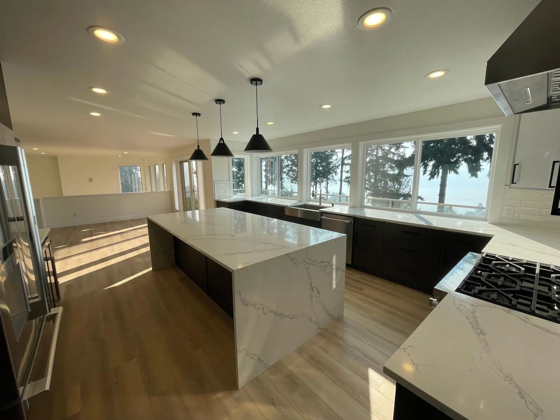 Modern kitchen with a large marble-topped island, dark cabinets, light wood floors, and floor-to-ceiling windows.