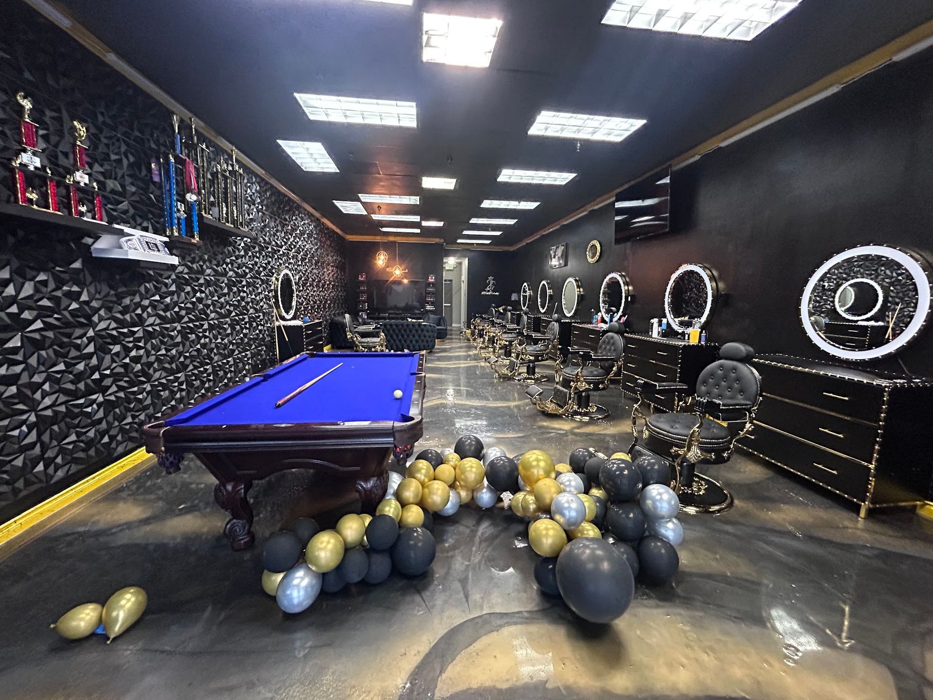 A decorated barbershop with pool table, balloons, and multiple barber stations. Dark walls, gold accents, and bright lights.