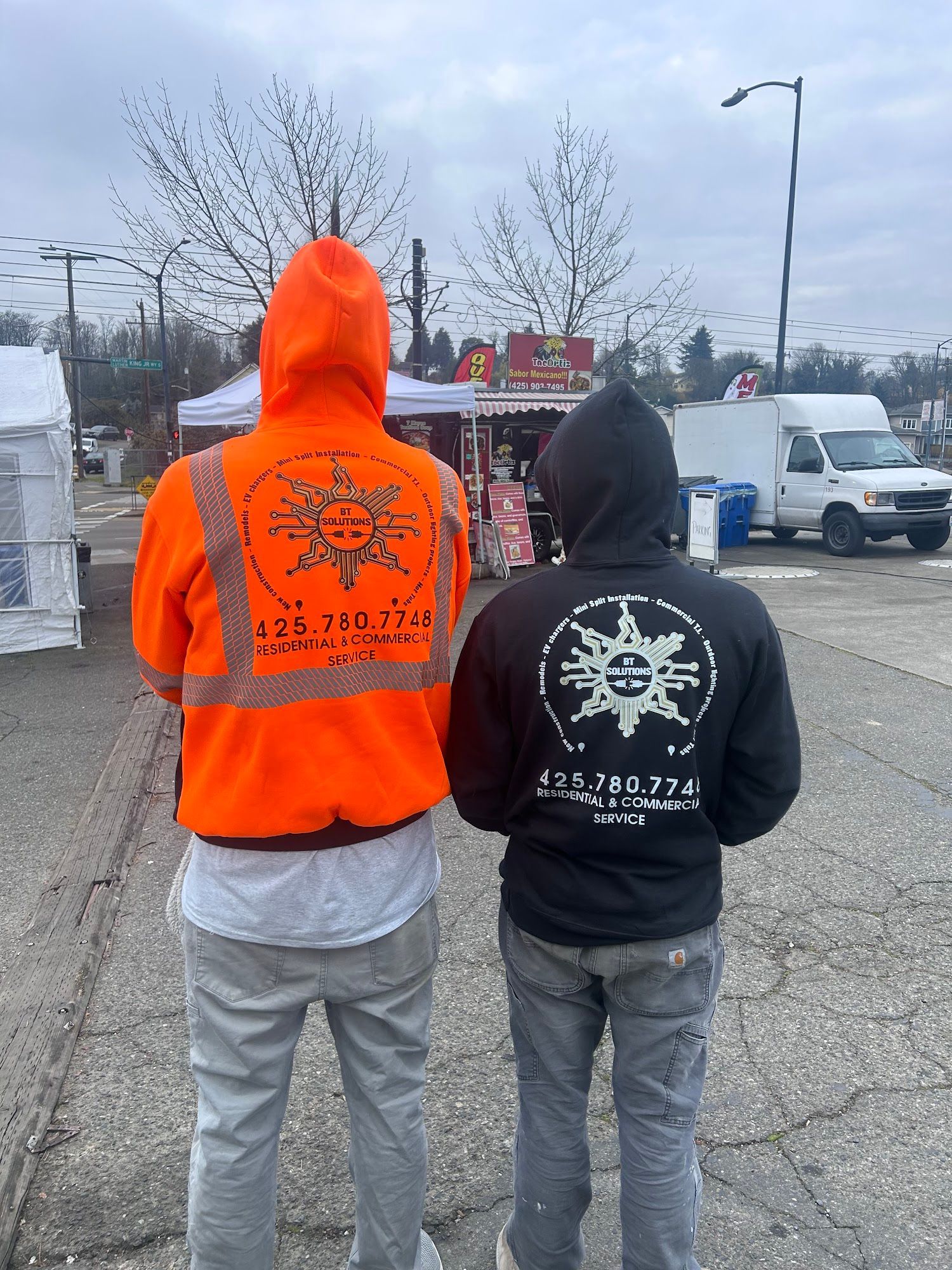 Two people wearing hooded jackets with a logo on the back, one orange, one black. They face away in a parking lot.