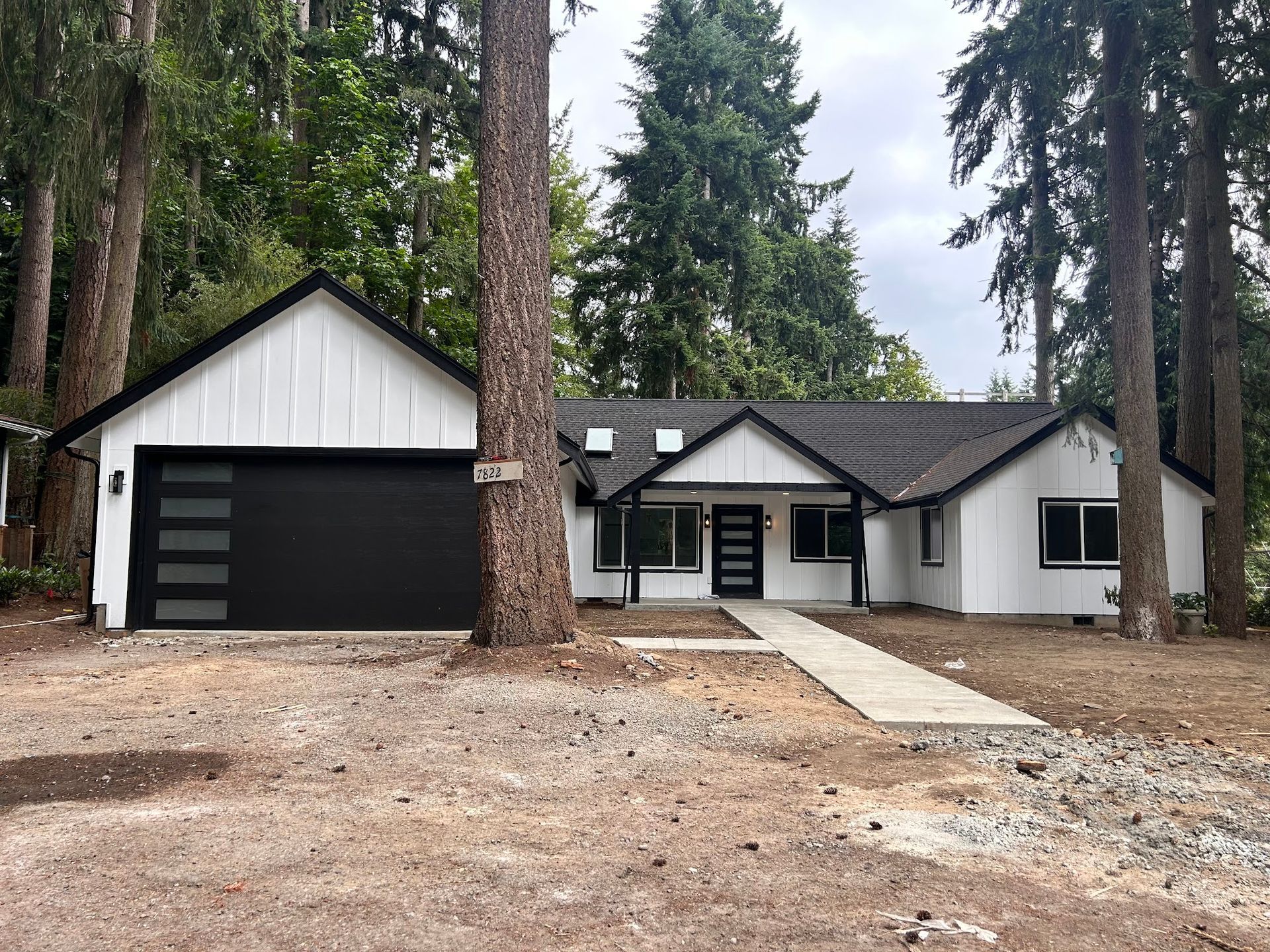 White house with black trim, garage door, and walkway; surrounded by trees.