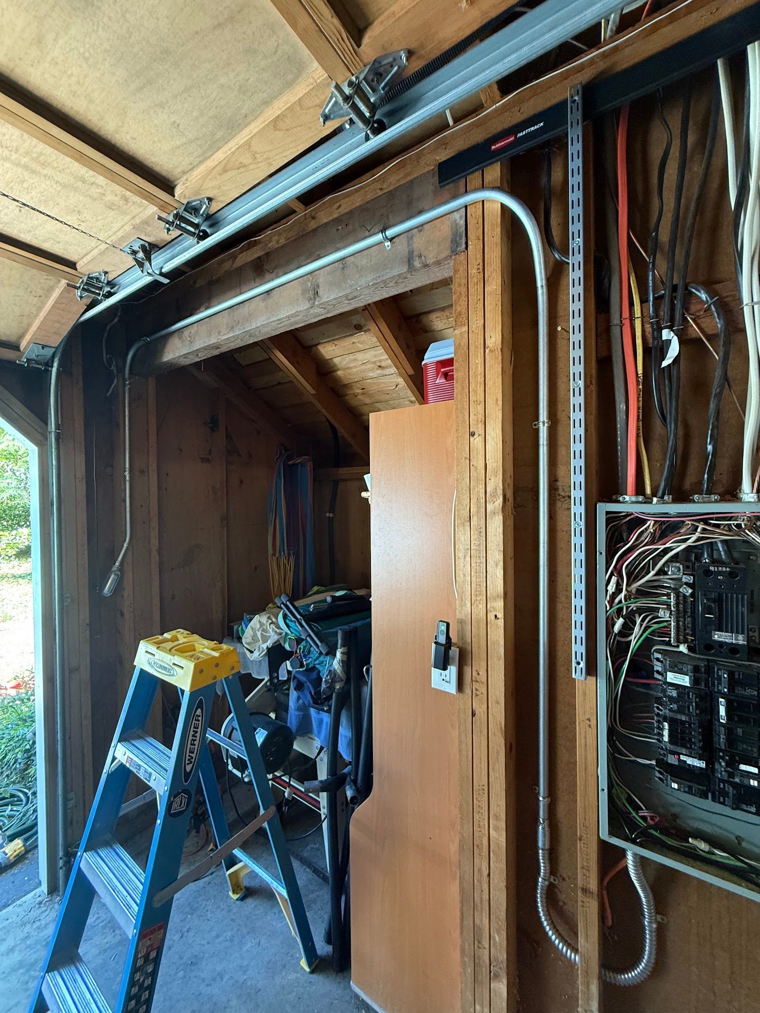 Electrical panel with conduit and wires in a wooden shed-like structure. A ladder leans nearby.