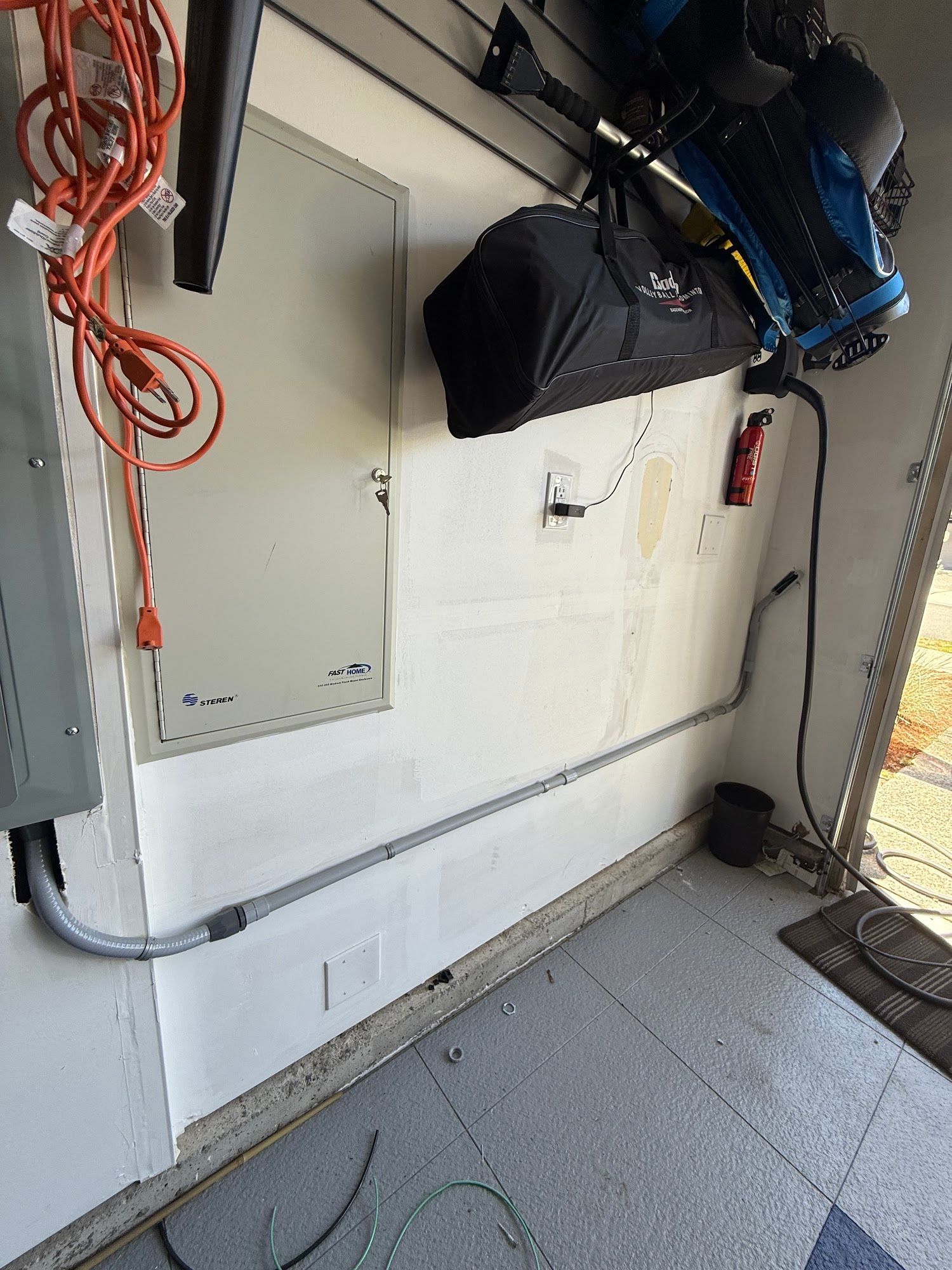Electrical panel in garage with conduit running along the wall. Extension cord, equipment, and a door are visible.