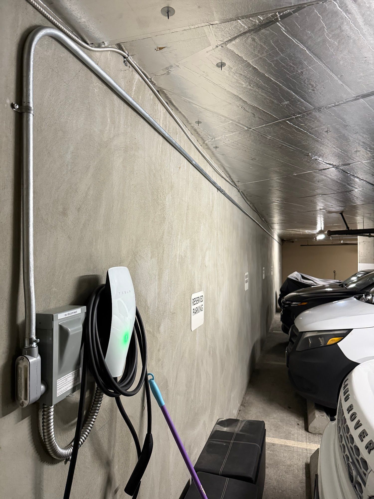 Electric vehicle charger in a parking garage. Conduit runs along concrete wall and ceiling. Cars parked in the background.
