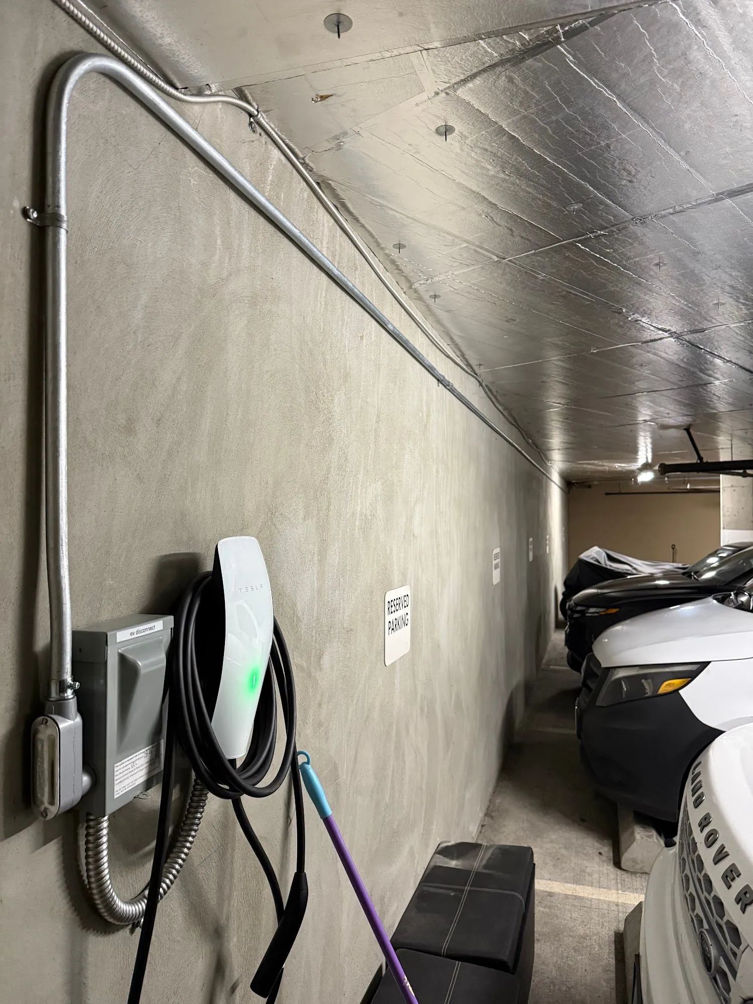 Electric vehicle charger mounted on a concrete wall in a parking garage, connected to metal conduit piping.