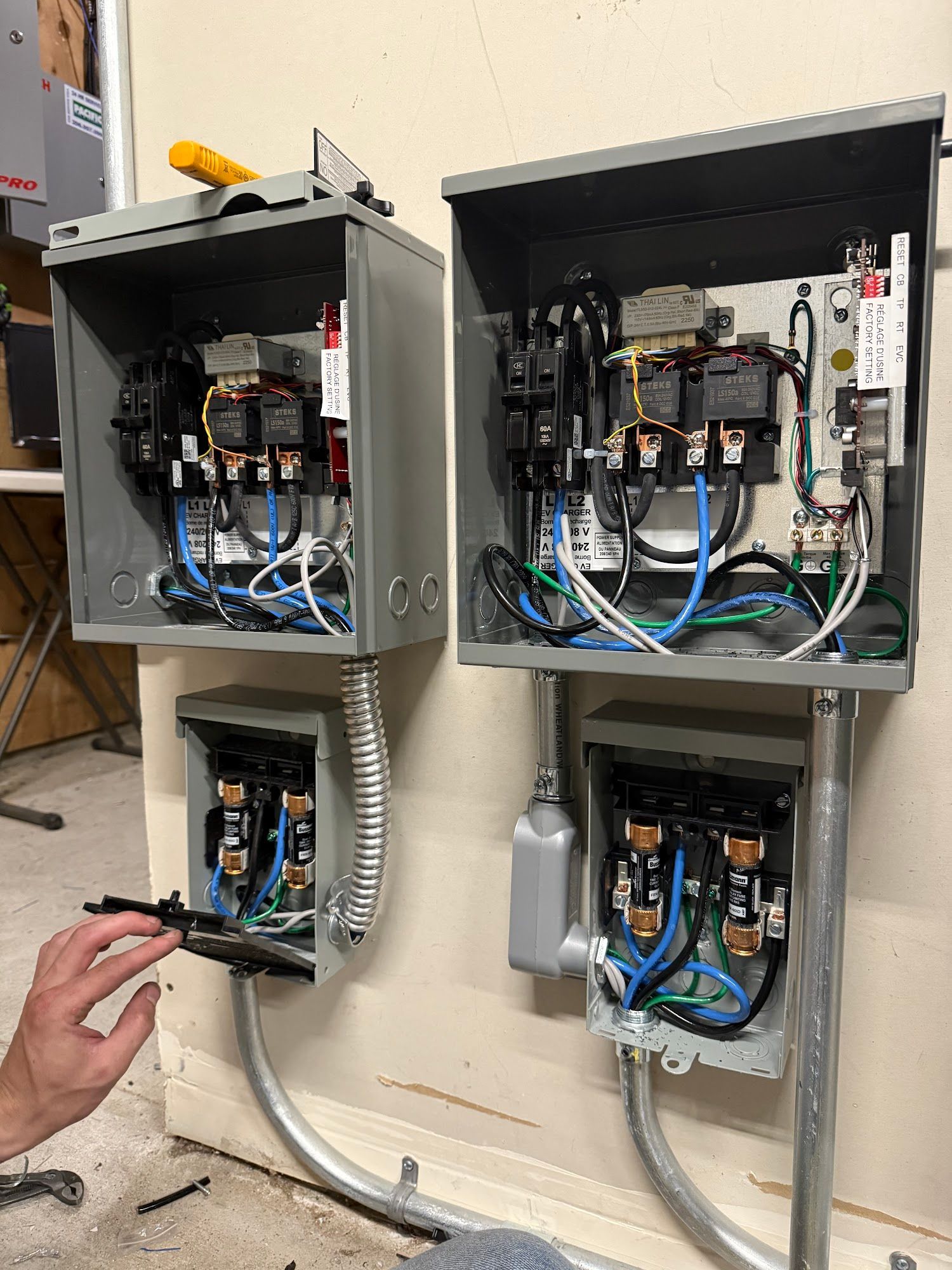 Electrical control boxes mounted on a wall; exposed wiring and components visible. Hand working on lower left box.