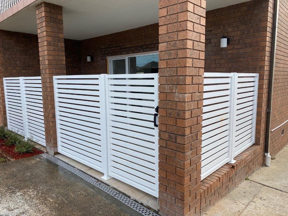 A White Fence Is Sitting on The Side of A Brick Building — Lifestyle Aluminium Fabrication In Albion Park Rail, NSW