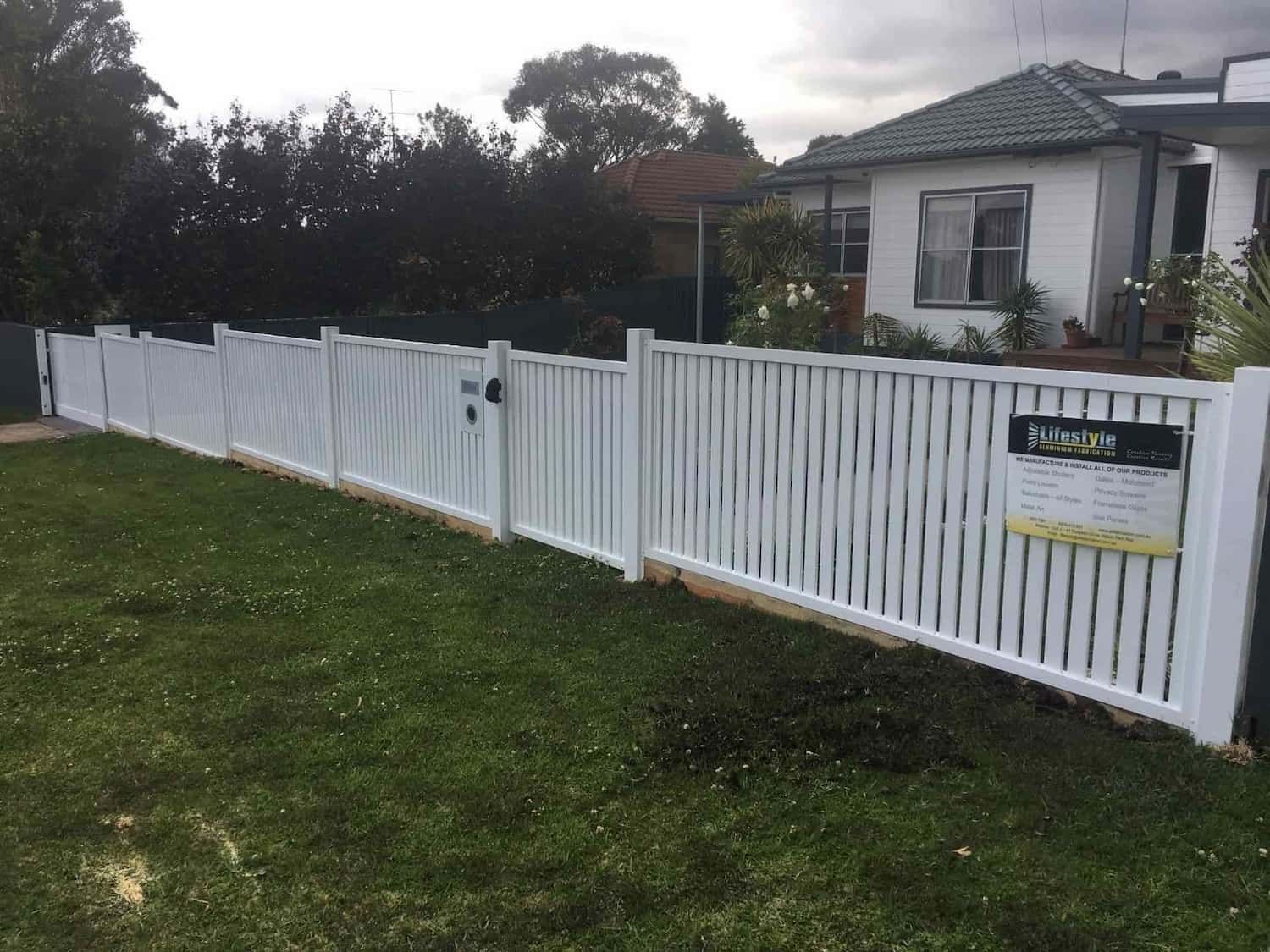 A White Fence Surrounds a Lush Green Yard in Front of A House — Lifestyle Aluminium Fabrication In Dapto, NSW