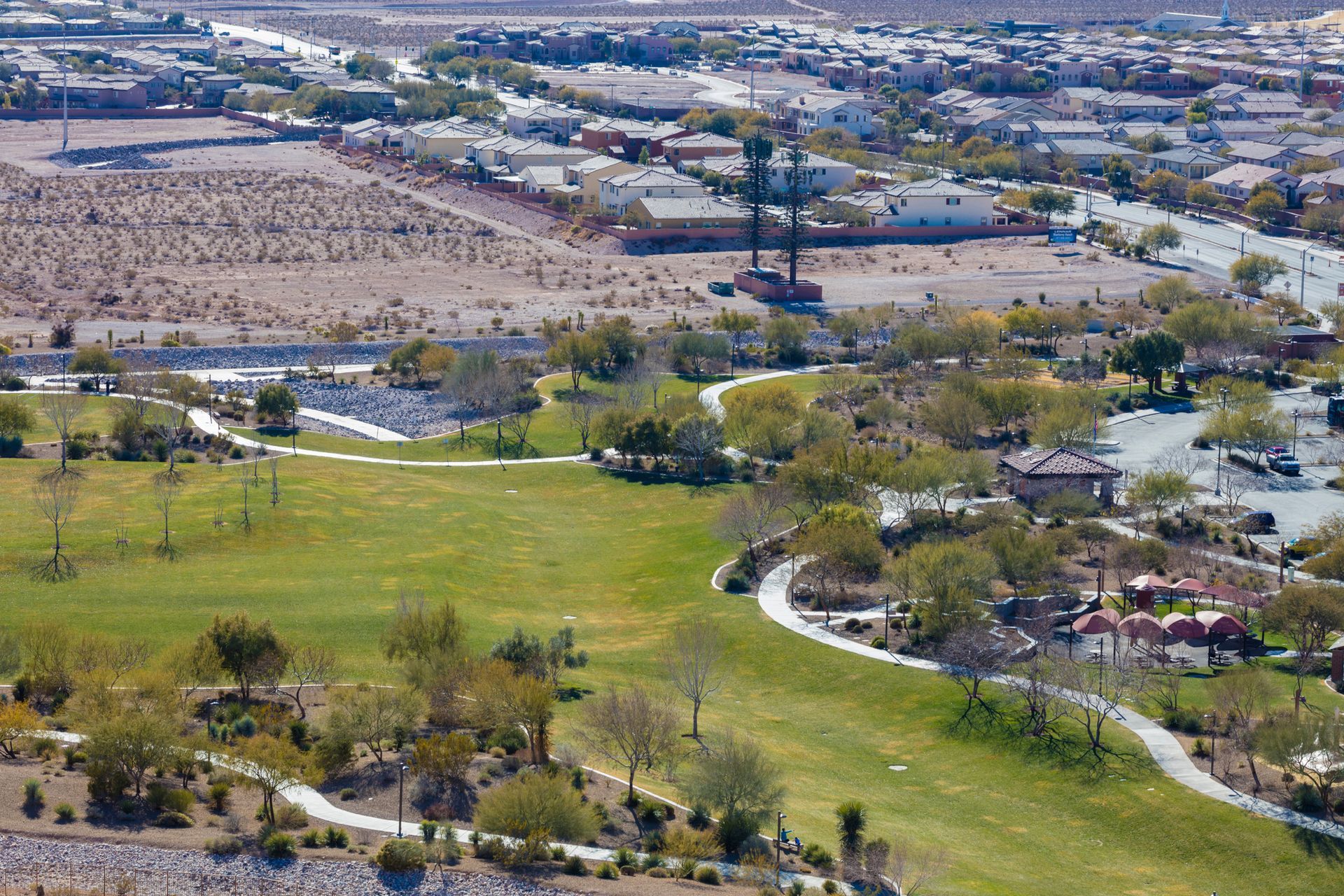 Green park with walking paths and trees, leading toward buildings in a desert landscape Green park with walking paths and trees, leading toward buildings in a desert landscape