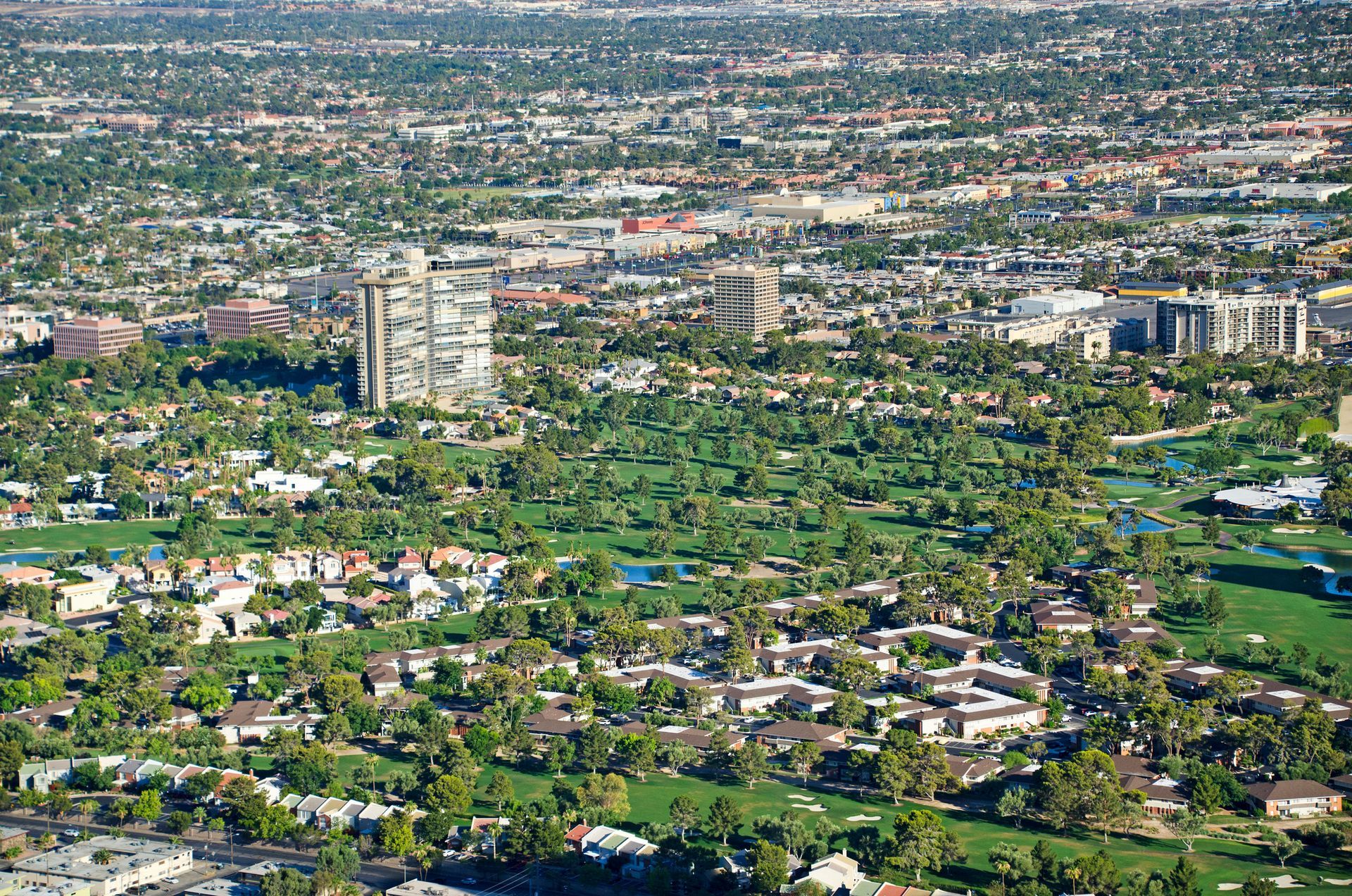 Aerial view of a city with a golf course, buildings, and many trees under a sunny sky