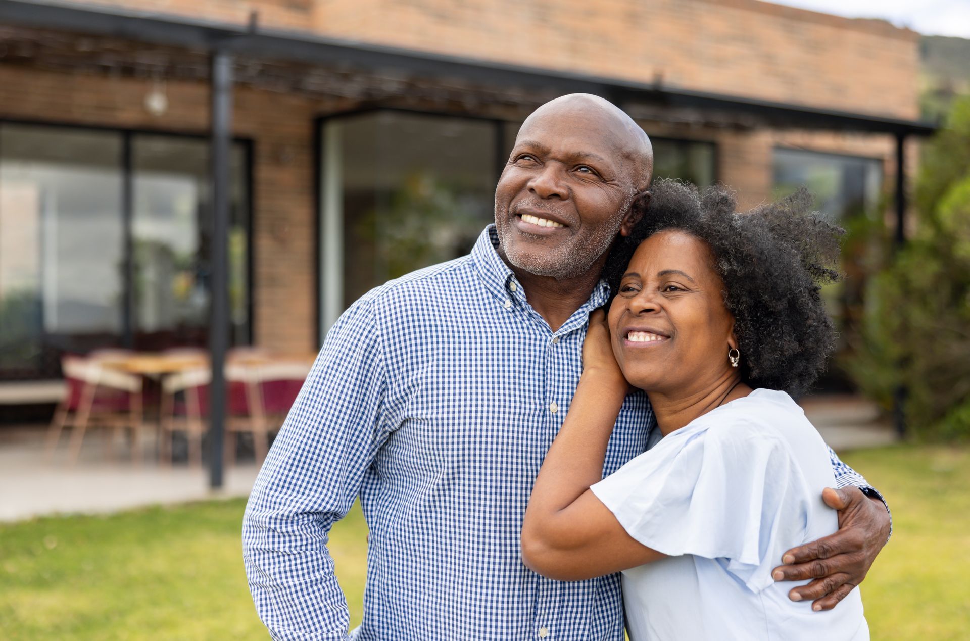Smiling elderly Black couple embracing outdoors, near a house with green lawn.