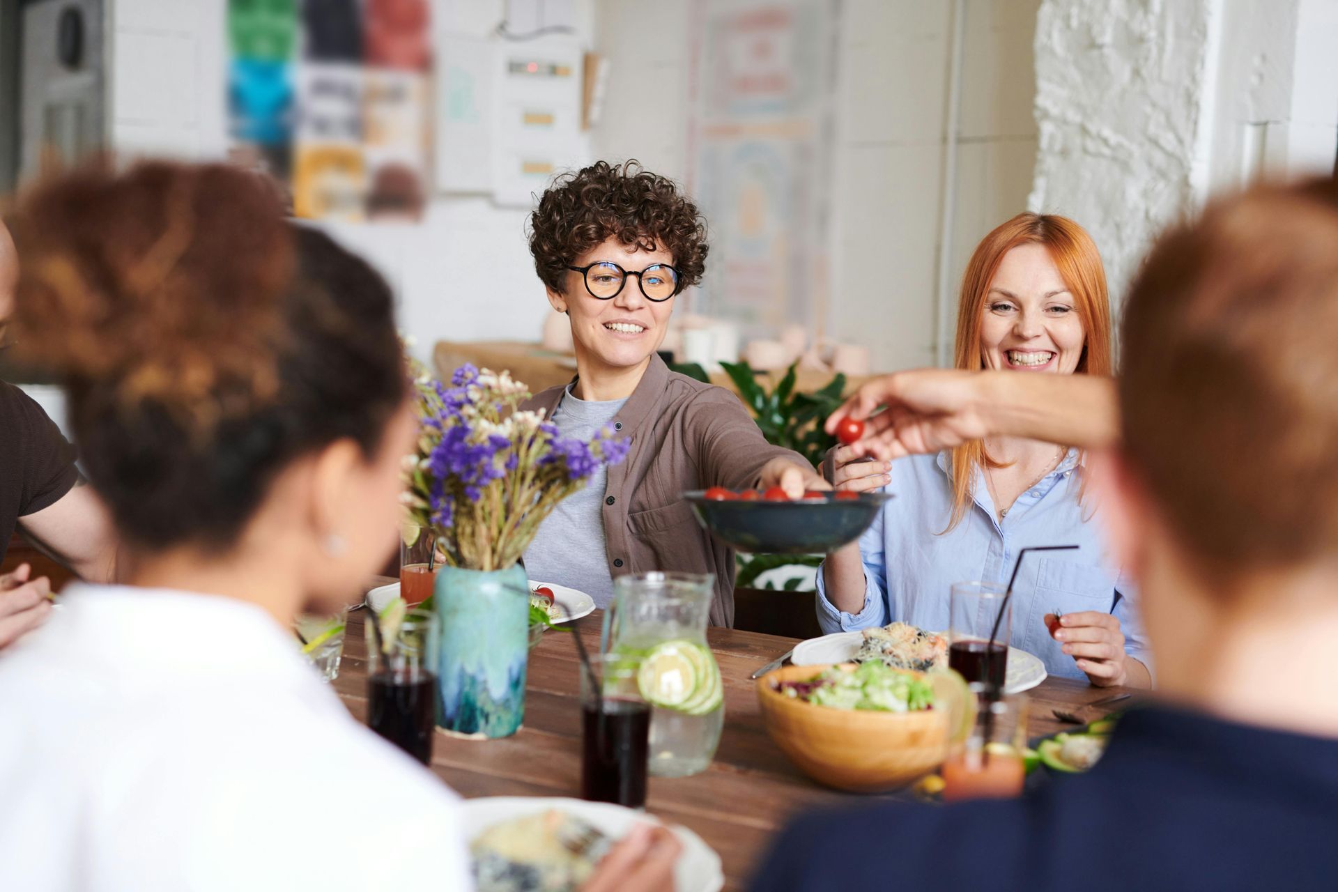 Friends at a table, sharing food, smiling. One woman holds a bowl of tomatoes, another smiles.