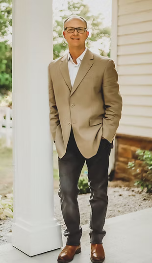 Man in glasses, tan blazer, and jeans, standing by a white column.
