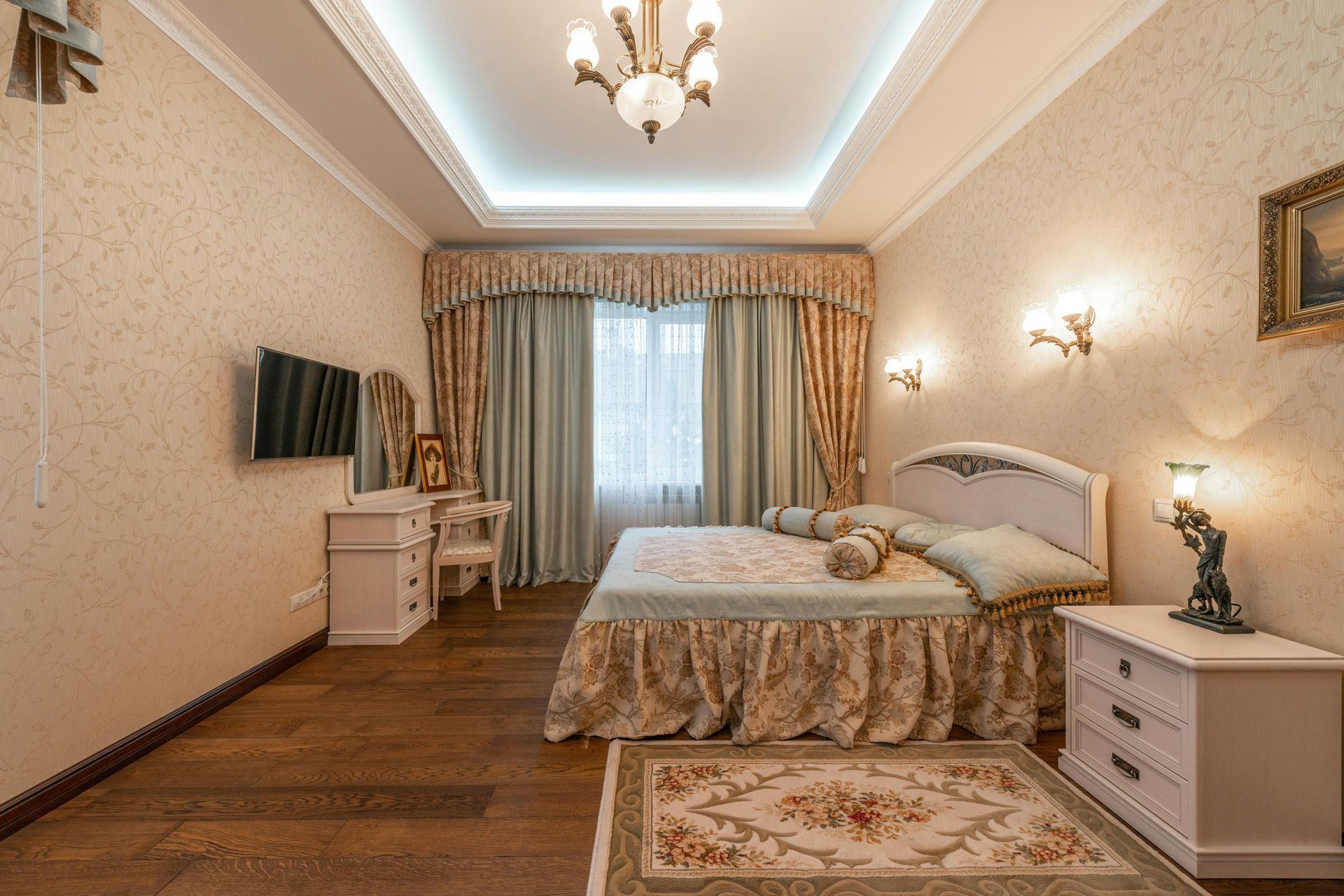 A bedroom with a large bed , dresser , nightstand , television and chandelier.