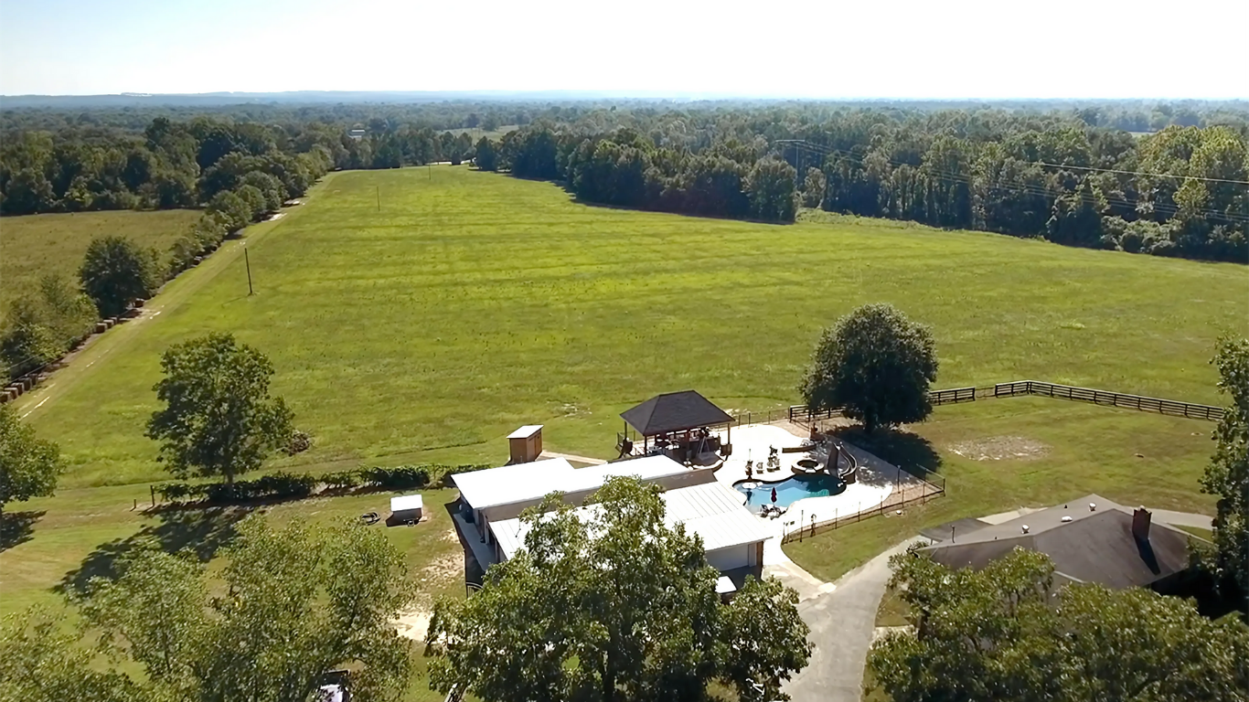 An aerial view of a house with a pool in the middle of a field surrounded by trees.