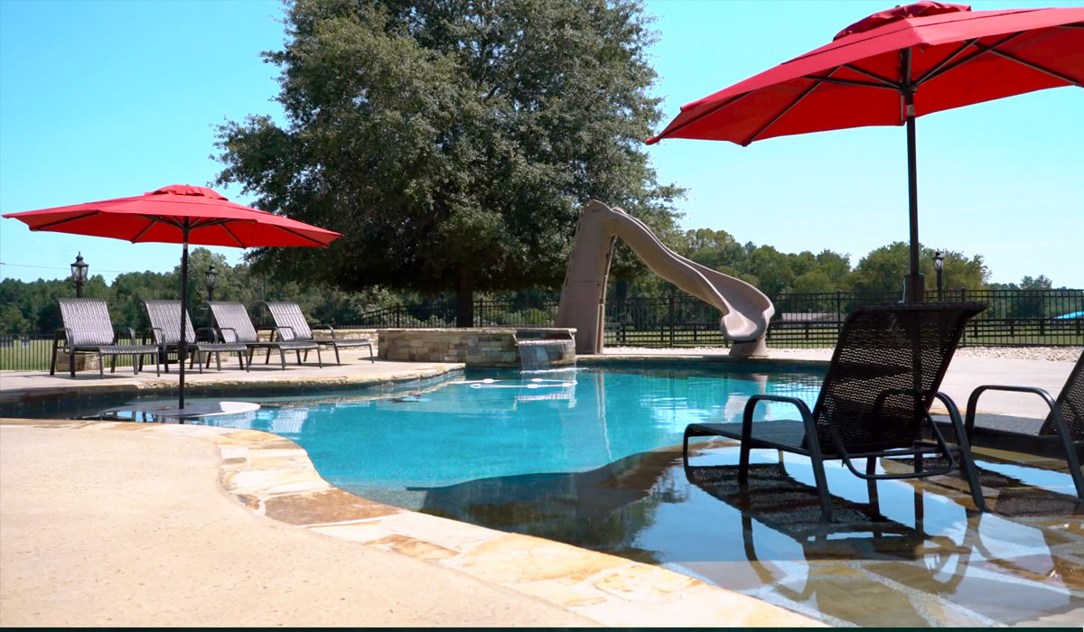 A swimming pool with a water slide, red umbrellas, and lounge chairs on a sunny day.