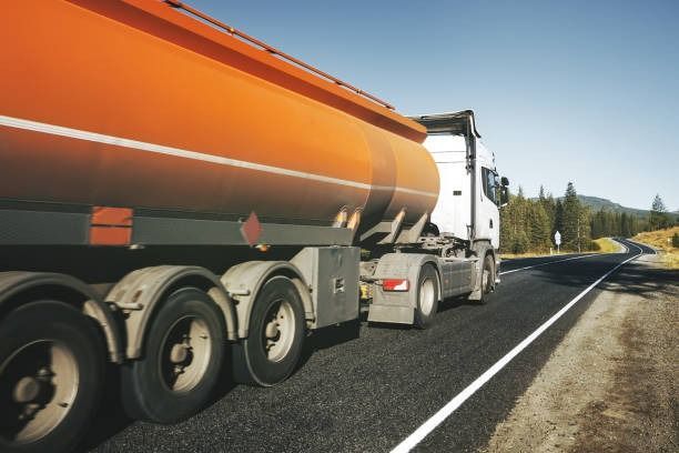 Camión cisterna de color naranja circulando por una carretera asfaltada, con montañas al fondo, día soleado.