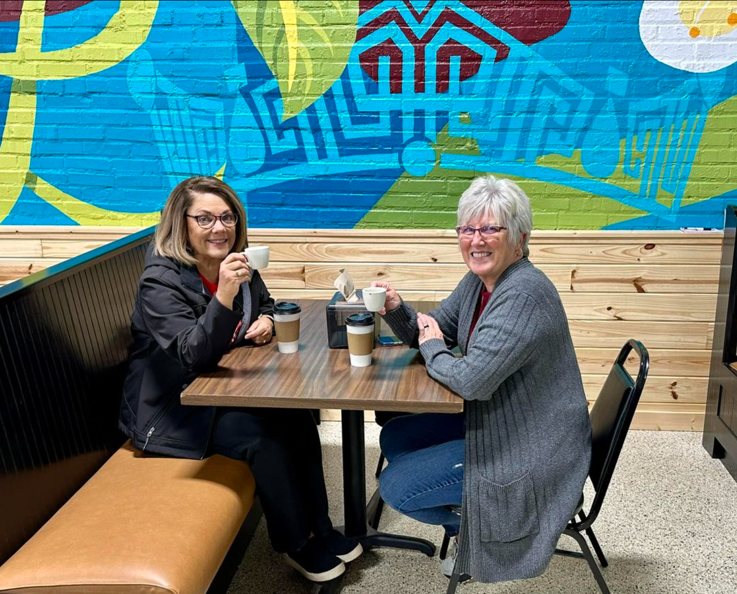 Two women are sitting at a table in front of a mural.