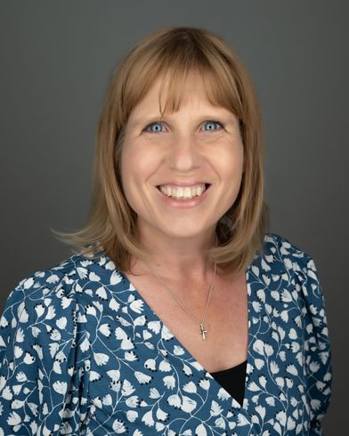 Woman with blonde hair and blue eyes, smiling, wearing a blue floral patterned top, against a gray backdrop.
