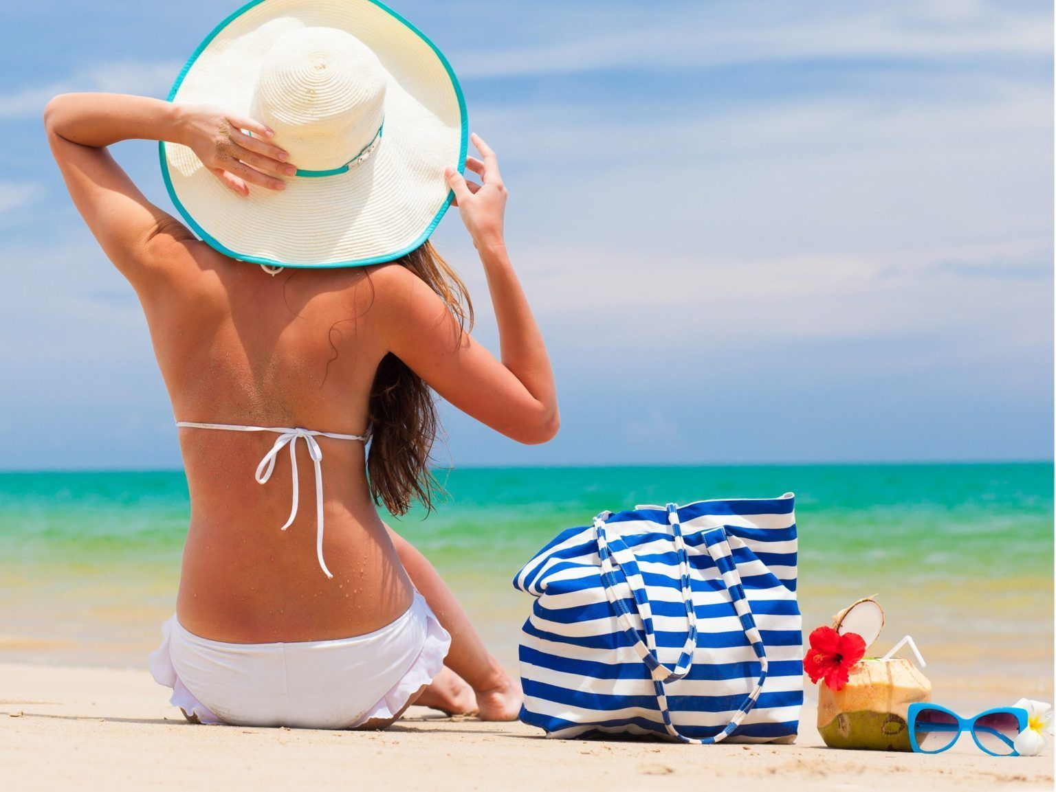 A woman is sitting on the beach wearing a hat and sunglasses.