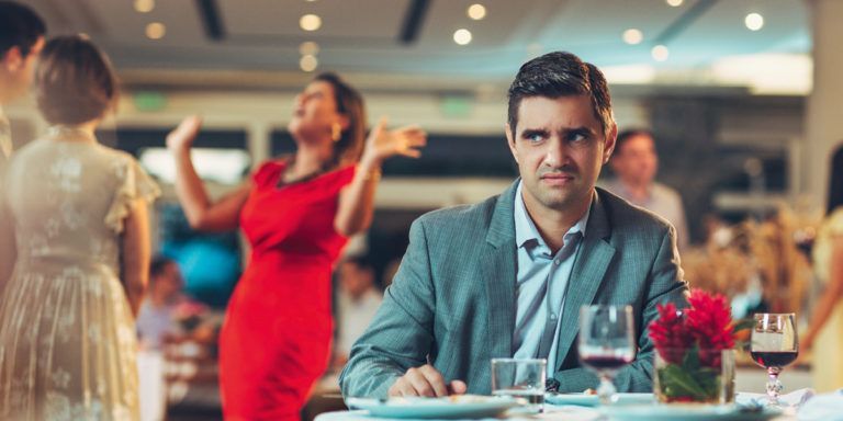 A man is sitting at a table in a restaurant with a woman in a red dress dancing in the background.