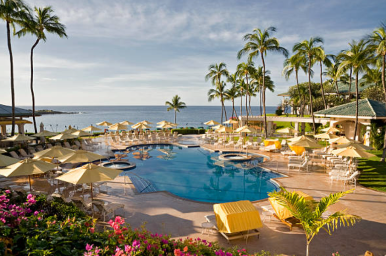 A large swimming pool surrounded by umbrellas and chairs with a view of the ocean.