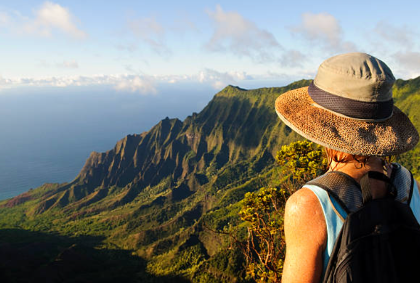 A woman in a hat is standing on top of a mountain looking at the ocean.