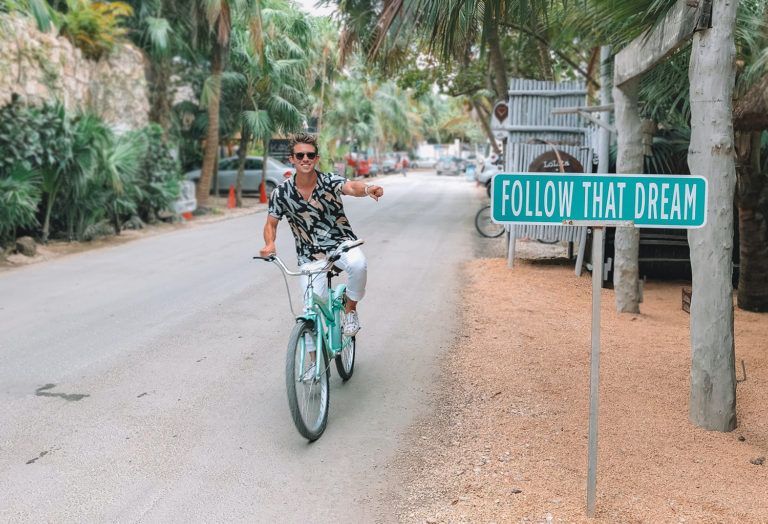 A man is riding a bike down a street next to a sign that says follow that dream.