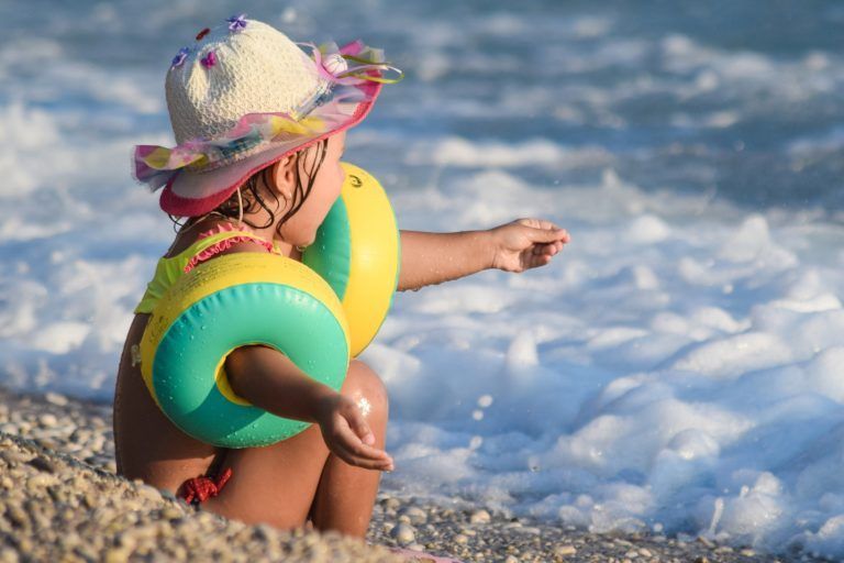 A little girl wearing a hat and life preservers is sitting on the beach.