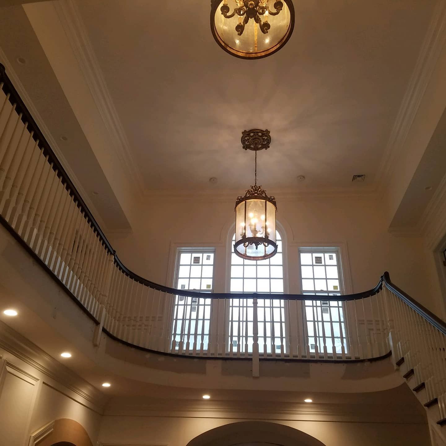 Interior view of a staircase with a curved banister and multiple windows. Two ornate light fixtures hang from the ceiling.