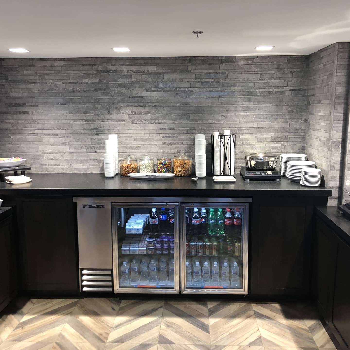 Bar with beverages: coolers, snacks, coffee machine, cups, on a counter with brick wall.
