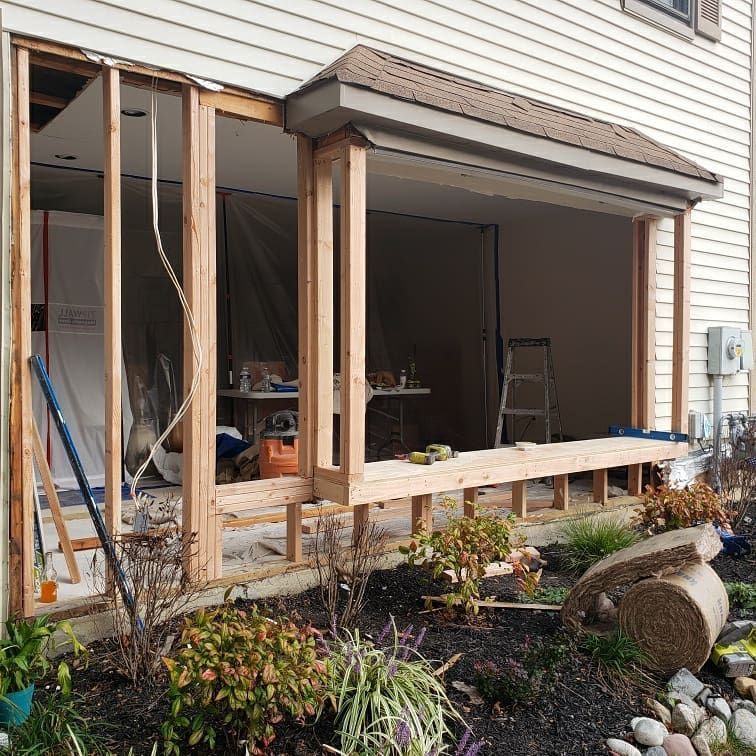 Exterior wall under construction, exposed wood framing, covered porch area, garden with shrubs and plants.