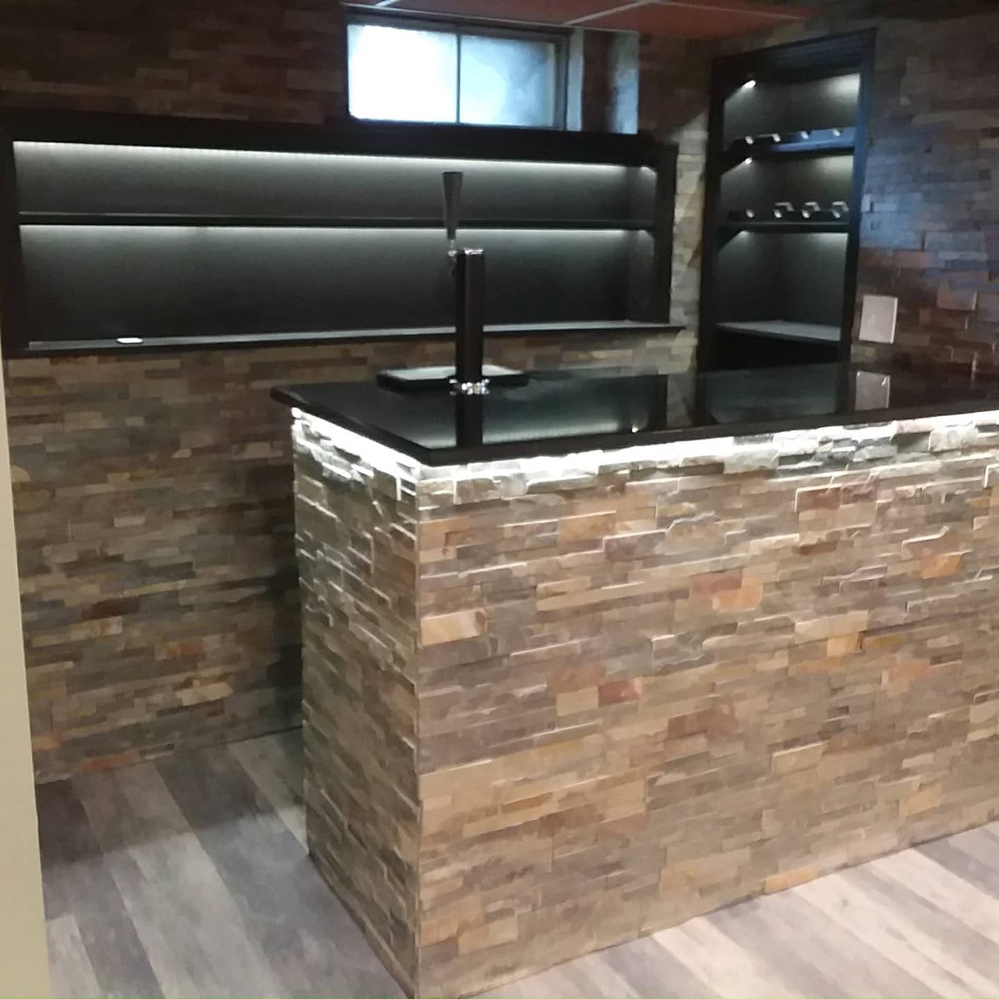 Basement bar with stone facade, black countertop, shelving, and built-in lighting.