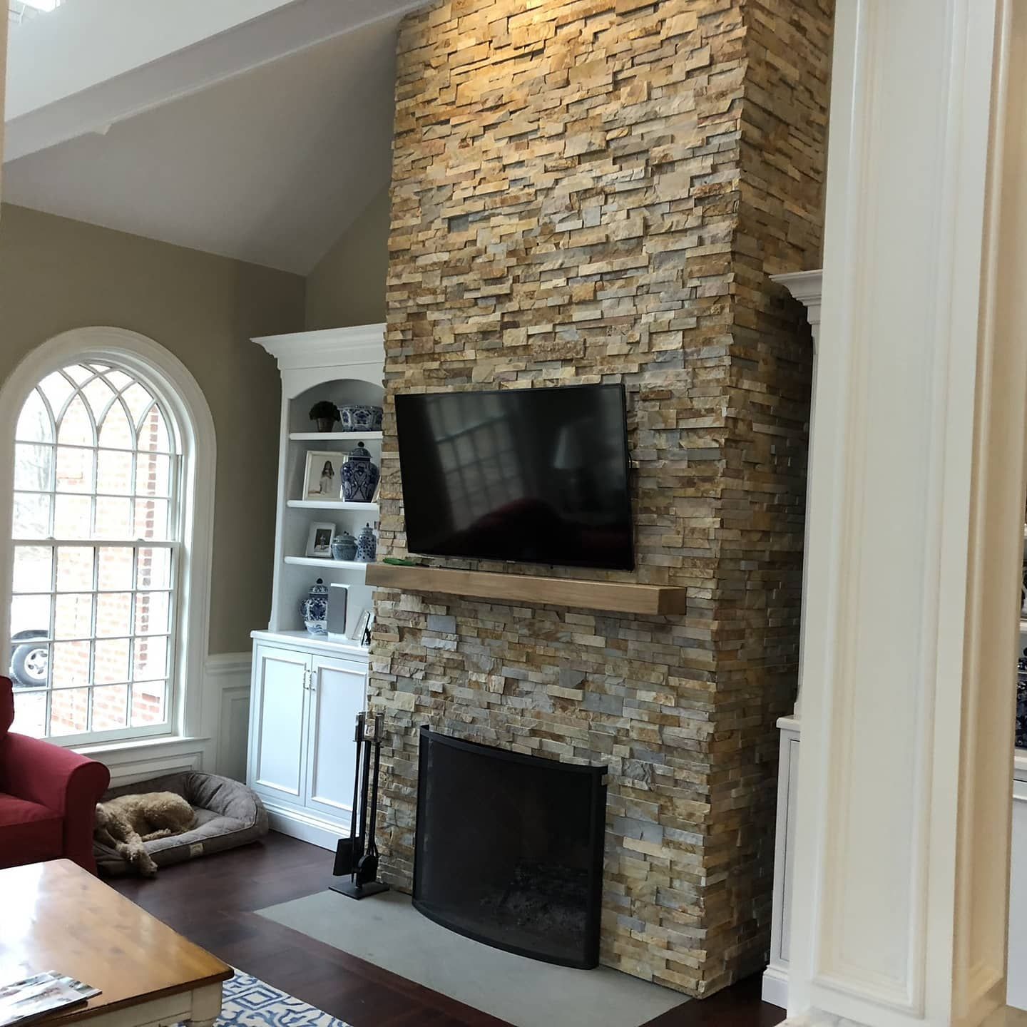 Living room with stone fireplace, TV, built-in shelves, arched window, red chair, and dog bed.