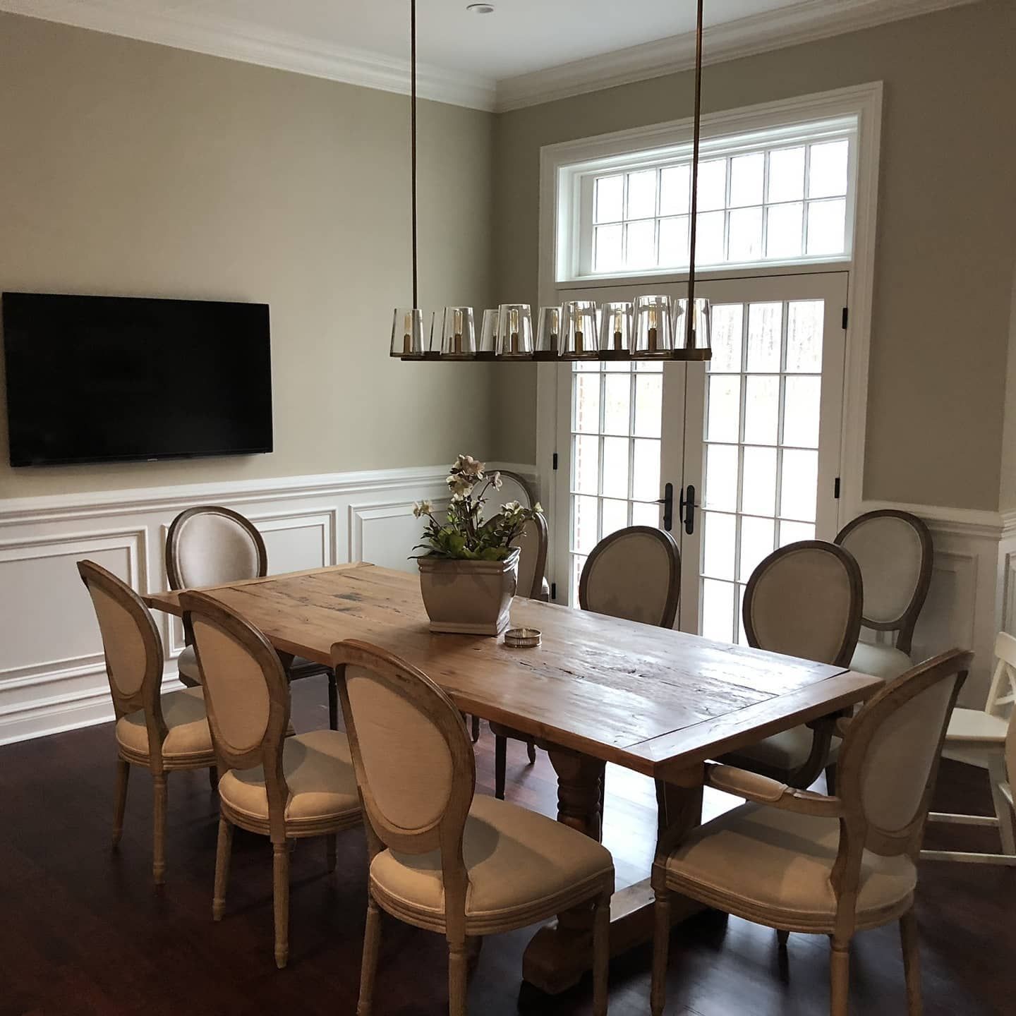 Dining room with wooden table, eight upholstered chairs, and a chandelier. A TV is on the wall.