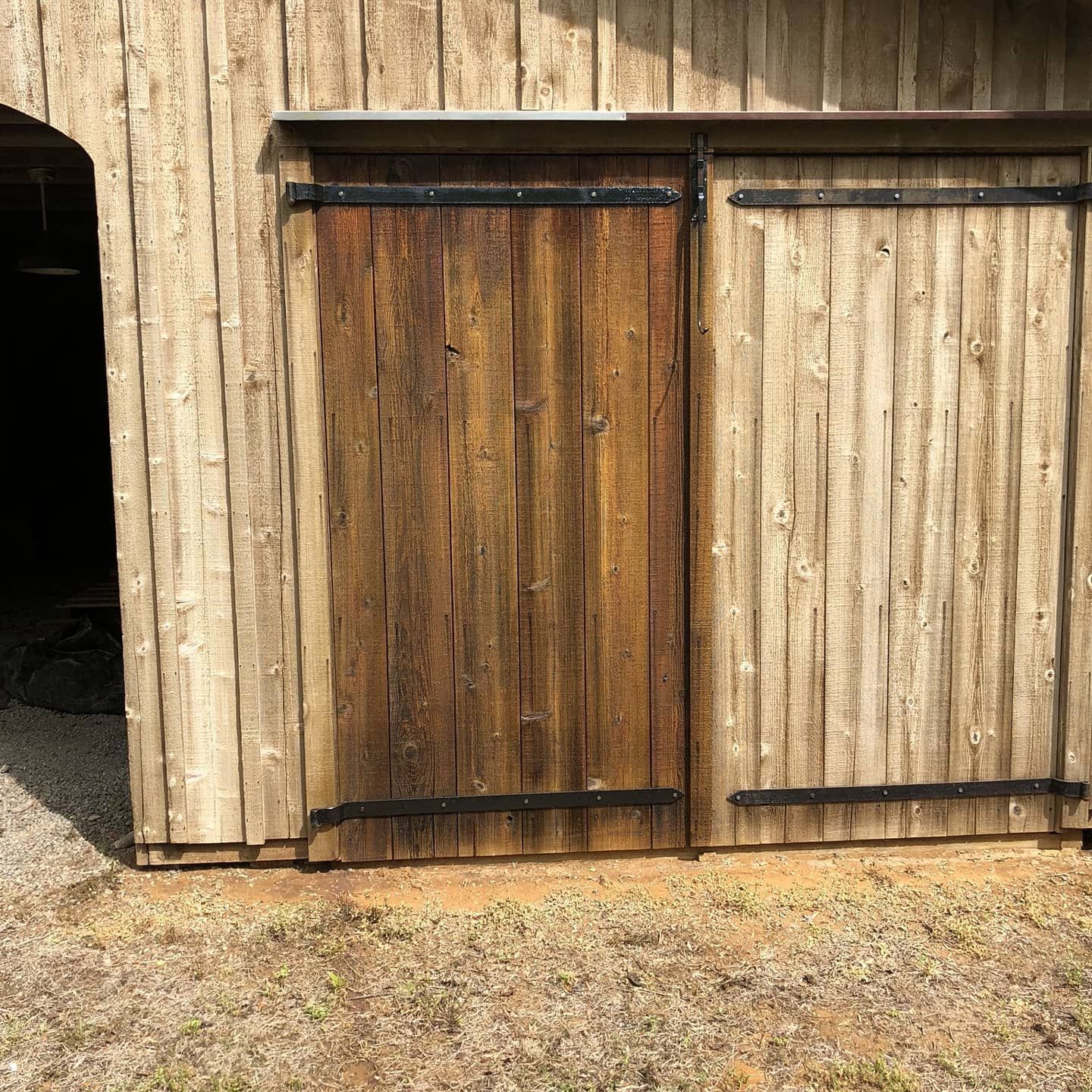 Wooden barn doors, one open, with dark metal hinges and trim. Brown, weathered wood.