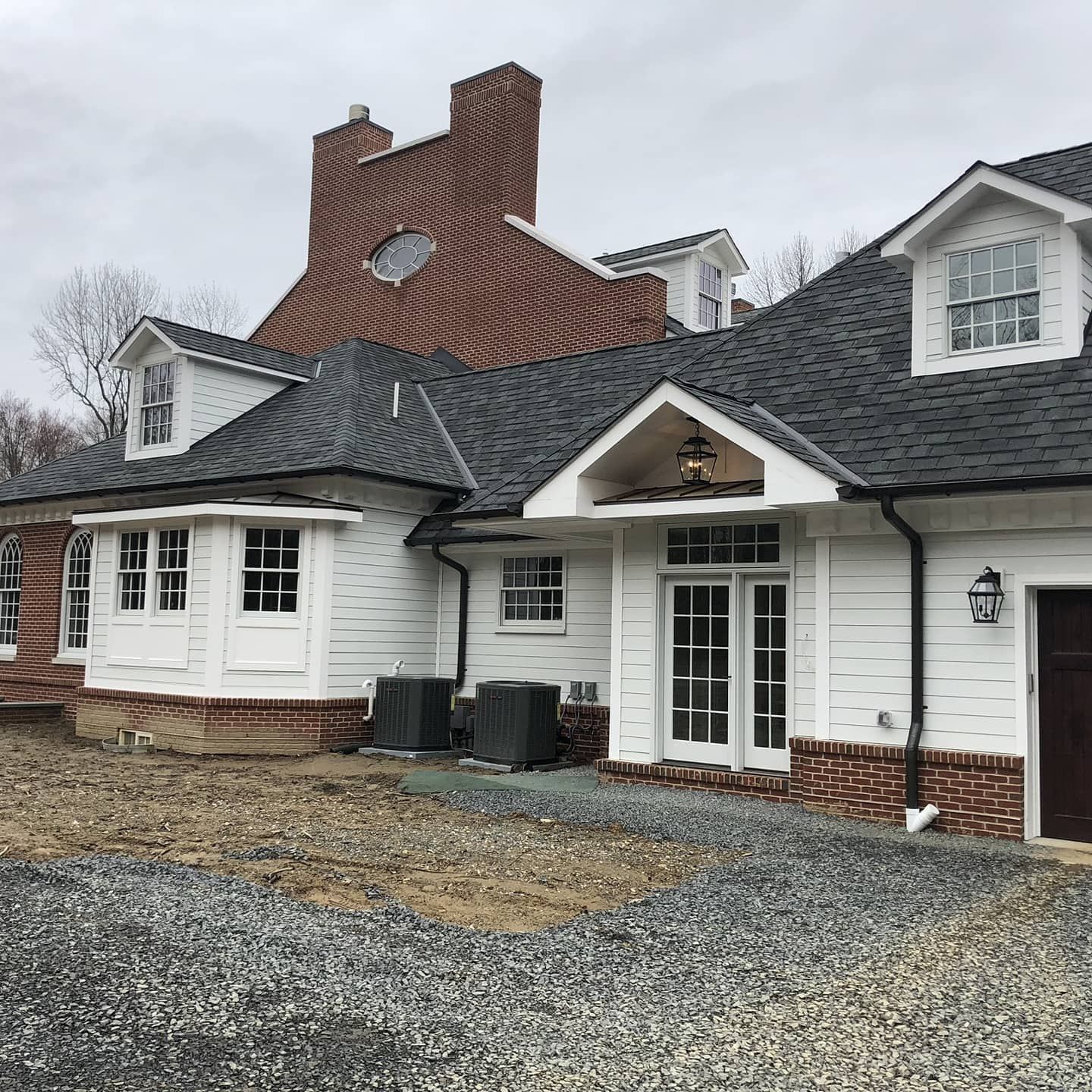White house with brick accents, gray roof, dormers, and chimney on a gravel lot.