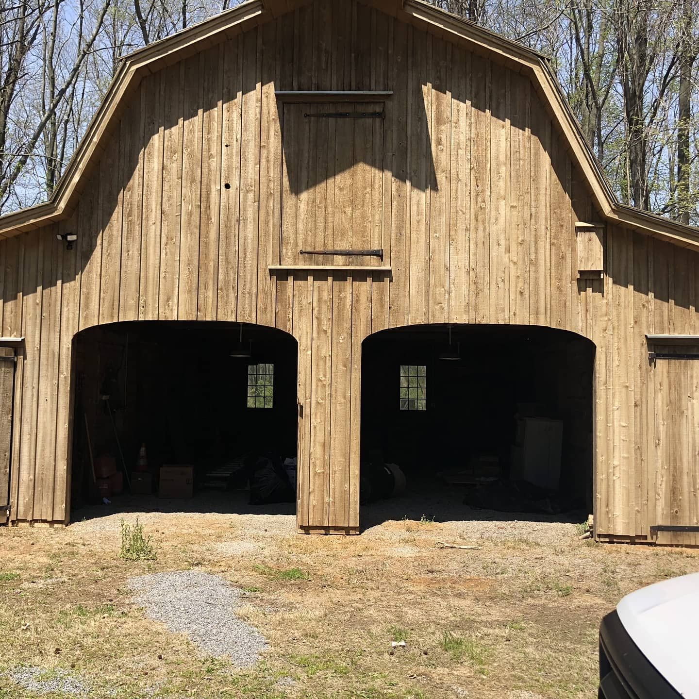 Wooden barn with two arched openings; gravel ground; wooded background.