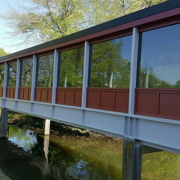 Covered bridge over water; red and brown siding, glass windows, blue sky reflection.