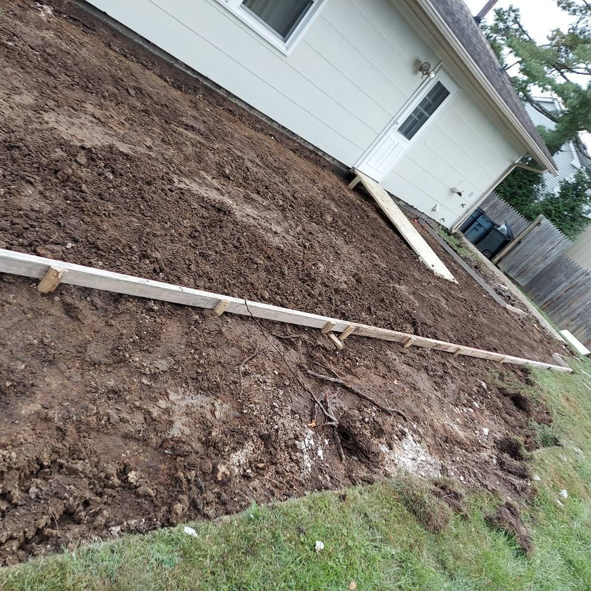 Dirt area next to a white house, prepared for landscaping. Wooden boards frame the space; grass on the edge.