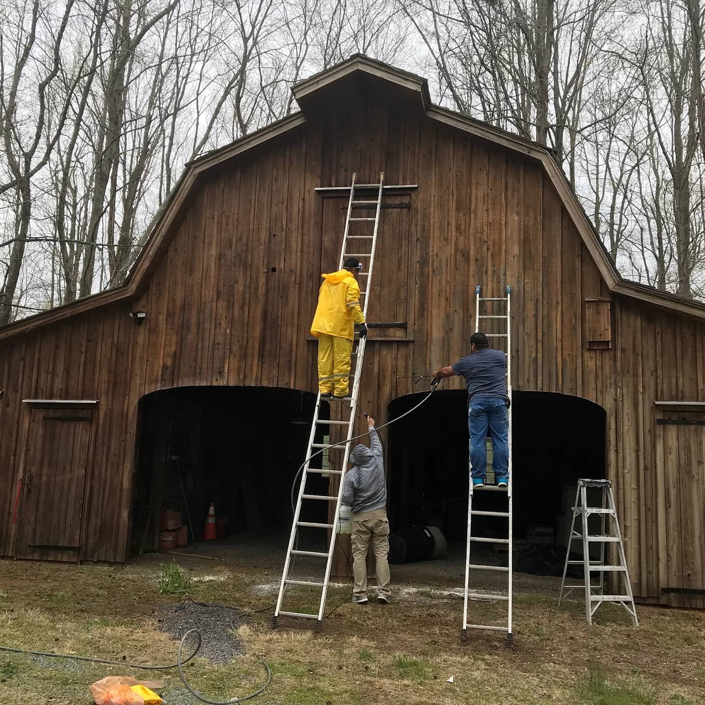 Three people on ladders working on a brown barn exterior. One wears a yellow jacket.